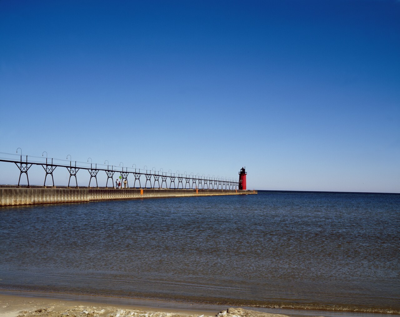 The red South Haven South Pierhead Light at the end of its catwalk, on calm Lake Michigan under a clear blue sky